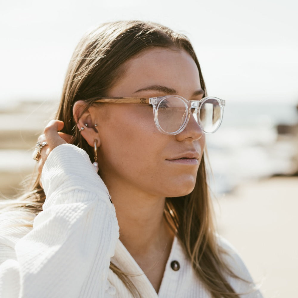 Woman wearing SOEK Wineglass Bay Blue Light Magnifying Readers with clear frames and bamboo arms, outdoors by the beach.