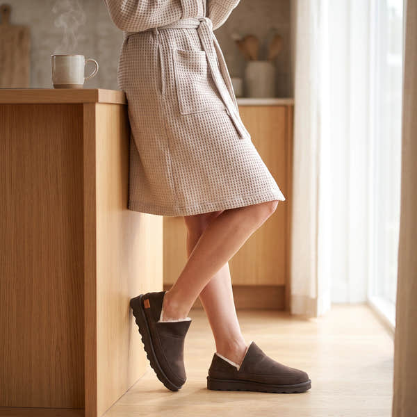 Strive Alaska chocolate brown suede slippers, worn with a robe in a kitchen