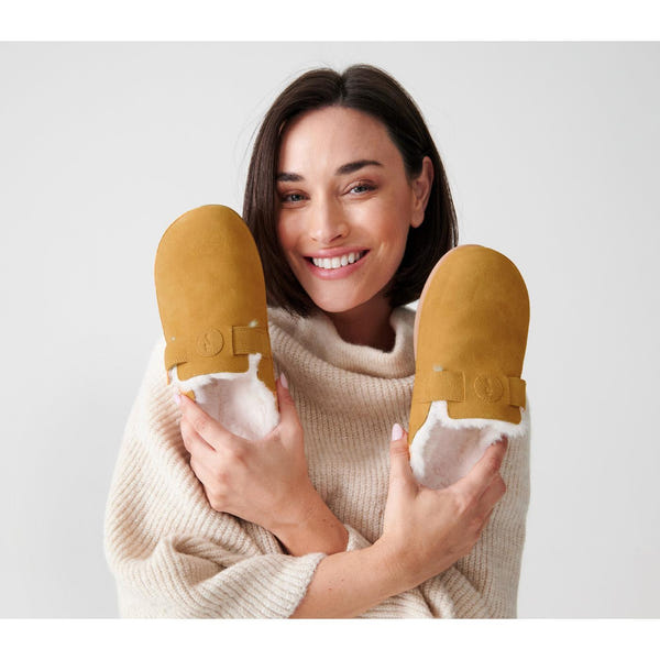 Woman smiling in beige sweater holding Strive Oslo Ladies Oak Leather Slip-On Slippers with arch support and faux fur lining.