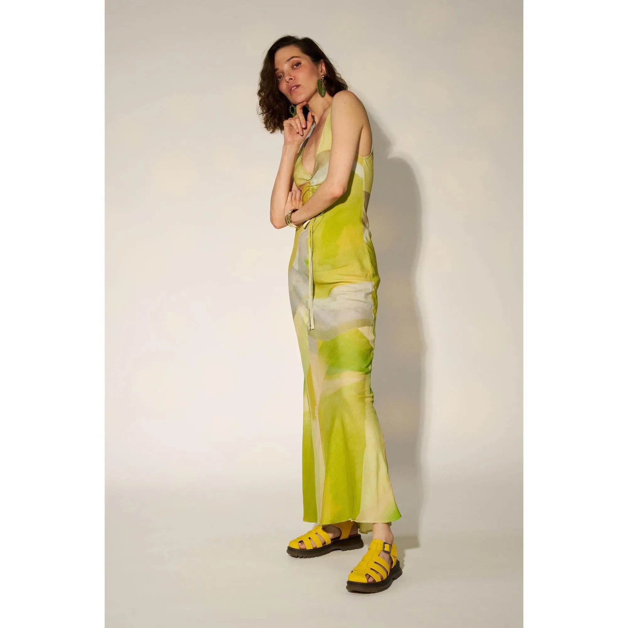 Woman in yellow tie-dye touch fastening sandals, green earrings, and matching yellow dress against plain background.