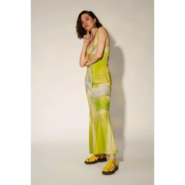 Woman in yellow tie-dye touch fastening sandals, green earrings, and matching yellow dress against plain background.