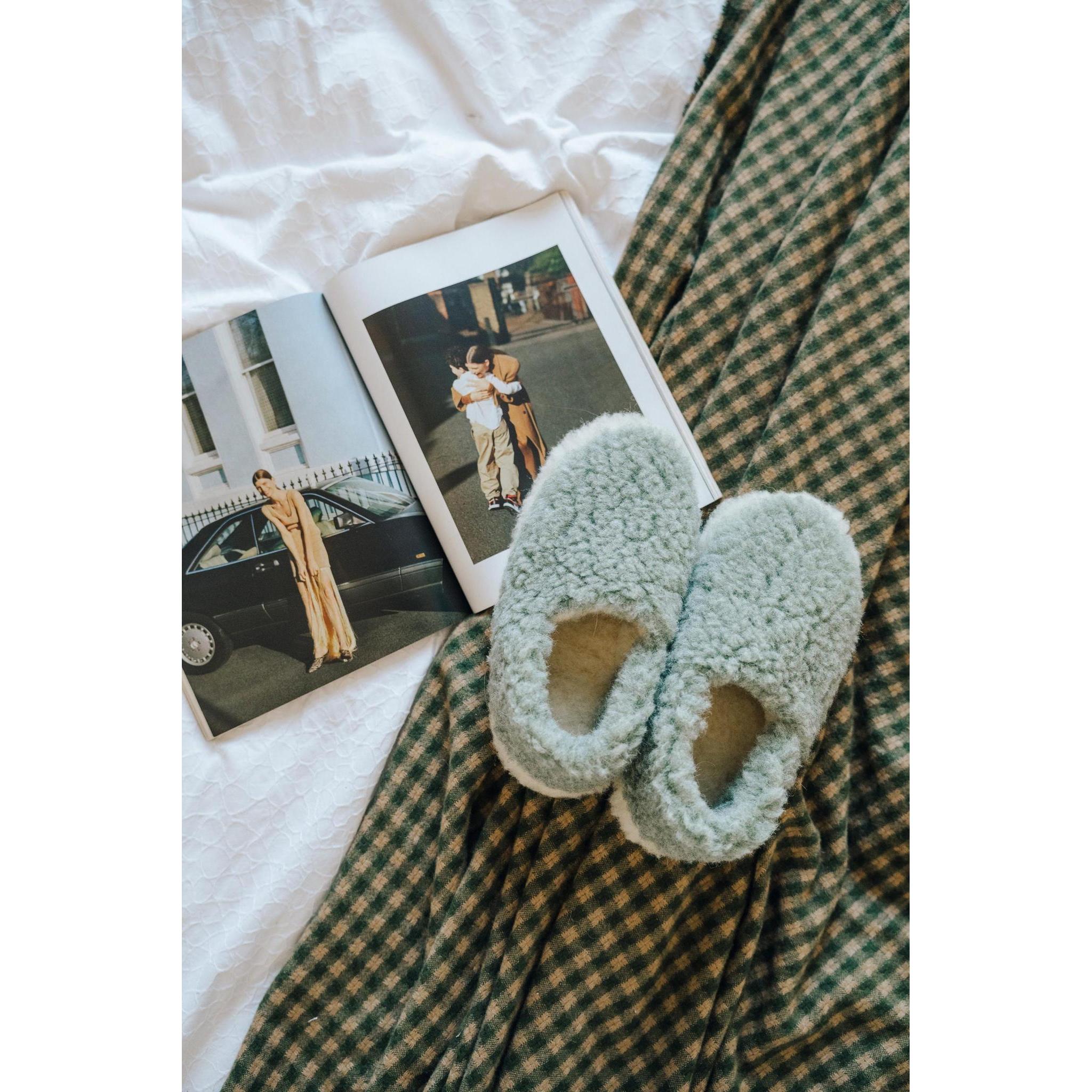 Yoko Full unisex Green wool slippers styled on a bed with a magazine, overhead lifestyle view.